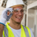 Male worker wearing safety glasses and a white hard hat. He is looking off camera and smiling. He is at a job site.