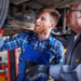 Mechanic working on area of car near the tire. He is wearing a flannel shirt, work gloves and blue overalls. He is being observed by another man who is also looking at the tire. That man is wearing blue coveralls, work gloves and glasses.