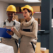 Two women working in a warehouse. They are looking at boxes and at a clipboard. Both are wearing overalls and yellow hard hats