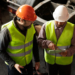 Two workers discussing a job. They are looking at each other. Both are wearing safety vests. One is wearing an orange hard hat, the other is wearing a white hard hat. There is manufacturing equipment in the back ground.